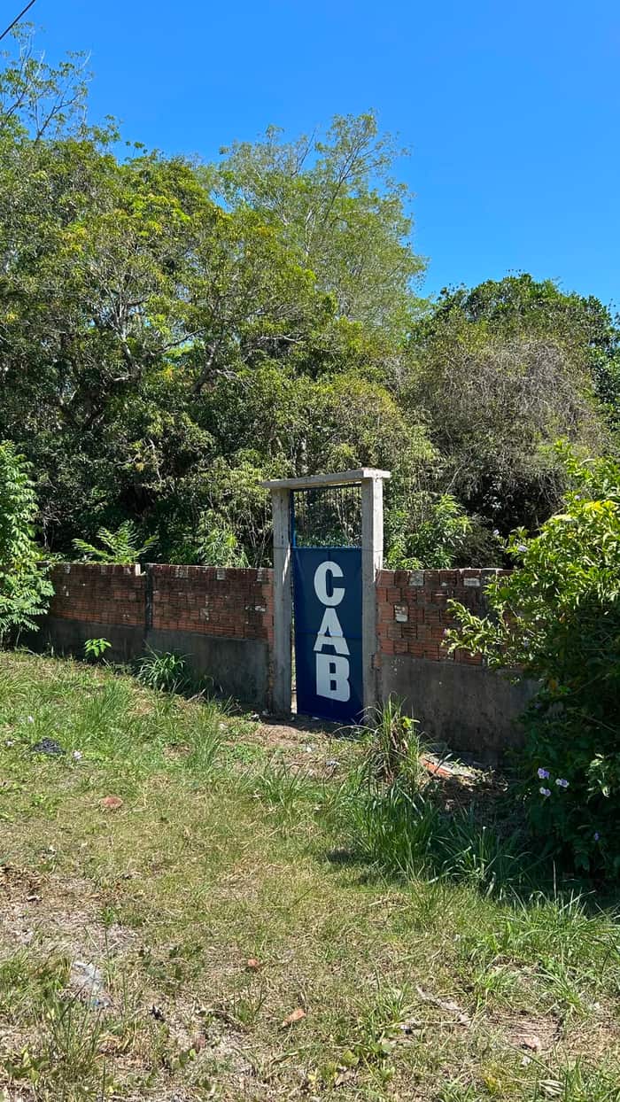 Muro de tijolos acompanhando o terreno gramado, com o portão de pedestres CAB ao centro e mata ao fundo
